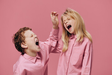 a boy and a girl of school age in pink clothes stand on a pink background and fight fiercely with...