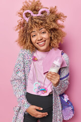 Positive curly haired young expectant woman touches belly poses with baby items smiles gladfully going to become mother soon wears warm clothes isolated over pink background. Pregnancy concept
