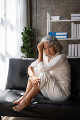 Sad depressed middle aged woman at home sitting on the couch, looking down and touching her forehead, loneliness and pain concept.