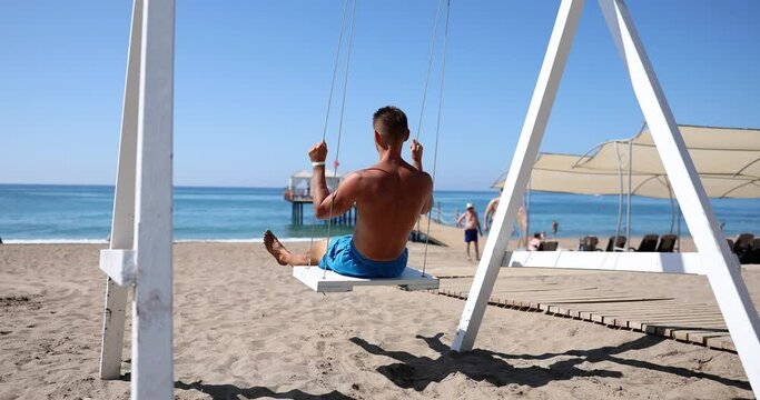 Male tourist traveler freelancer blogger is resting on a swing on sandy beach against backdrop of beautiful sea island