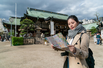 Asian Japanese woman visitor holding and reading route map on Osaka Tenmangu Shrine plaza in japan with the main hall at background on a cloudy day in spring.