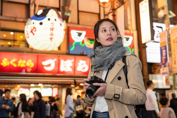 curious Asian Chinese female photographer holding camera and looking into space while exploring Shinsaibashisuji shopping area street in Osaka japan at night