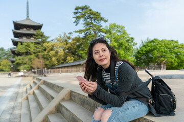 happy asian Japanese girl looking into distance enjoying breeze while using smartphone for online guide near kofuku-jiâs five storied pagoda in nara japan