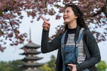 happy asian Japanese female tourist pointing at famous attraction in distance while visiting Kofuku-ji temple with five storied pagoda and sakura flowers at background in nara japan