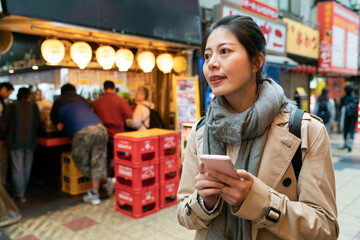 smiling asian korean girl visitor using phone gps to navigate while shopping at food district in Shinsekai area in Osaka Japan. she looks into space checking the right way