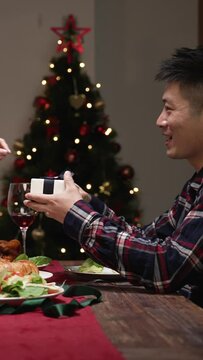 Vertical Screen: asian woman taking out Christmas gift from her back to surprise boyfriend who is feeling happy after opening box at dinner table on the festive holiday at home