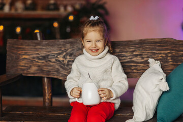 A cheerful baby is drinking hot chocolate on a bench by the fireplace. Christmas mood, Christmas...