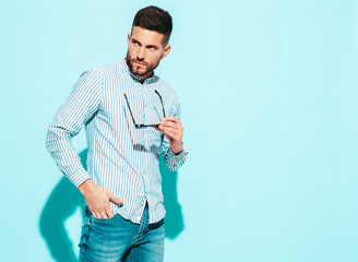 Fototapeta premium Portrait of handsome confident model. Sexy stylish man dressed in shirt and jeans. Fashion hipster male posing near blue wall in studio. Takes off sunglasses
