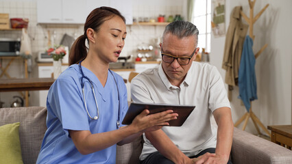 Asian female nurse using hand gesture while showing checkup results on tablet computer to elderly male patient at home. the man listens with worried face expression