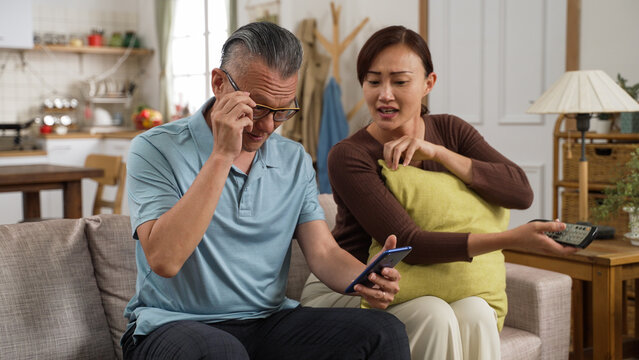 Adult Adult Daughter Watching Tv Helping Give Father To Take Reading Glasses While He Is Using Cellphone At Home. The Smiling Senior Male Free From Bad Vision After Wearing Eyeglasses