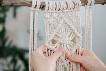 Hands working on a macrame piece suspended from a stick. Central part pattern is nicely done and square shaped. Blurred room in background.