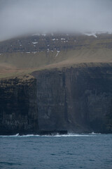 Fototapeta na wymiar Impressive steep cliffs and rocky coastline silhouette coast of Faroe Islands in Atlantic Ocean on stormy grey day with clouds, lighthouse and hills seen from cruiseship cruise ship liner with spray