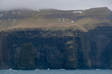 Impressive steep cliffs and rocky coastline silhouette coast of Faroe Islands in Atlantic Ocean on stormy grey day with clouds, lighthouse and hills seen from cruiseship cruise ship liner with spray