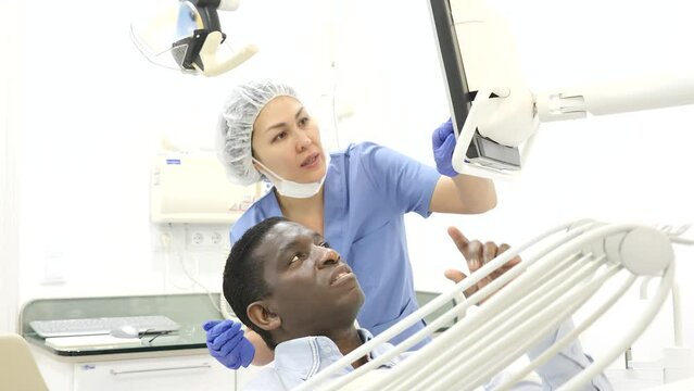 Confident asian female stomatologist showing teeth radiography result on computer screen to african american male patient, discussing medical treatment at clinic