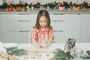 Little dark-haired girl 3 years old in red checkered shirt prepares gingerbread cookies from dough in white Christmas decorated kitchen with garland lights. Merry Christmas, Happy New Year