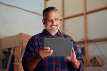 Happy mid adult man in checked shirt using digital tablet at woodworking factory