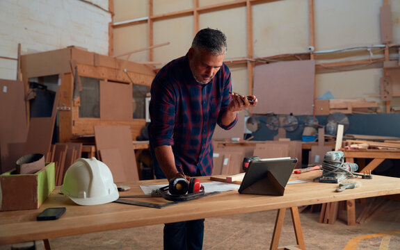 Mid Adult Man Talking On Mobile Phone While Writing On Paper In Woodworking Factory