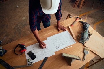 High angle view of mid adult man in safety helmet writing on paper near digital tablet in woodworking factory