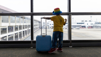 Fototapeta na wymiar Child with suitcase at airport terminal waiting for departure looking out the window. Child and suitcase at the airport, indoors and waiting for going to travel. 
