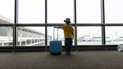 Child with suitcase at airport terminal waiting for departure looking out the window. Child and suitcase at the airport, indoors and waiting for going to travel.	