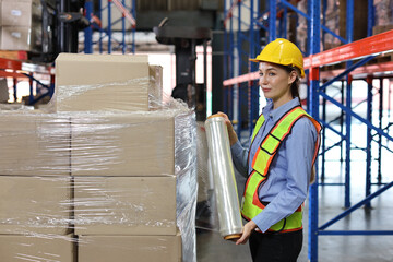 Caucasian warehouse worker woman with hardhat and reflective jackets wrapping boxes in stretch film parcel on pallet while control stock and inventory in retail warehouse logistics distribution center