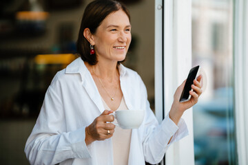 White mature woman drinking coffee and holding cellphone in cafe