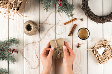 Female hands tie bow on gift box in festive package, top view. Spruce branches, cinnamon, cup of coffee and twine ball. Crafting present. Objects layout. Christmas or New Year holiday concept