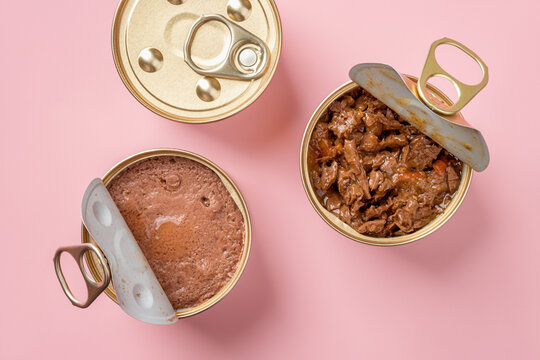 Wet cat food in open jars over pastel pink background. Wet pet feed in metal cans closeup. Canned meat pieces and soft pate for carnivore domestic animals. Сat food concept.