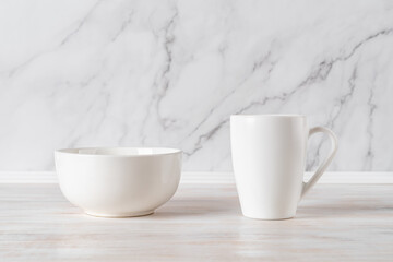 Bowl and mug on a table against marble wall. Blank ramekin and large cup mockup for food and drink design. Modern crockery and white tableware concept.