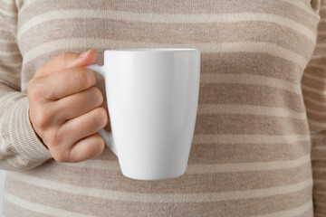 Woman hand holding white coffee mug. Female hand holds blank tea cup against striped wool sweater. Modern tableware, white crockery, hot beverage concept.