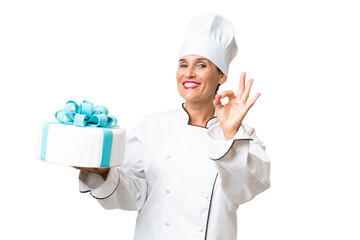 Middle-aged chef caucasian woman with a big cake over isolated background showing ok sign with...