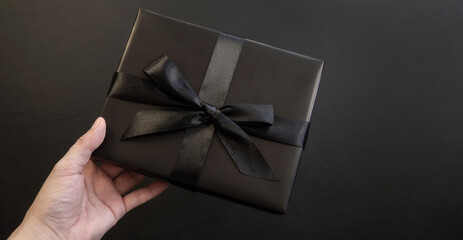 Gift box in hand wrapped in black paper with a black bow on a dark background. Holiday concept. Place for text or advertising. Christmas. Black Friday.
