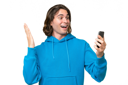 Young handsome man isolated on green chroma background looking at the camera while using the mobile with surprised expression