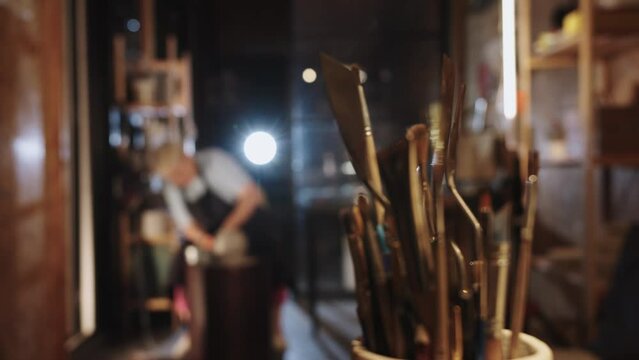 An elderly woman making a clay pot in the workshop - brushes on the foreground