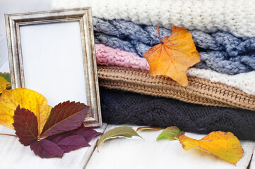 Hello autumn. A stack of knitted winter clothes, colorful autumn leaves and an old empty picture frame with copy space on a bright wooden table. Pile of knitted cosy warm jumpers.