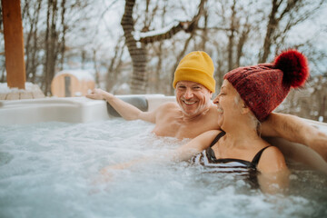 Senior couple in kintted cap enjoying together outdoor bathtub at their terrace during cold winter day.