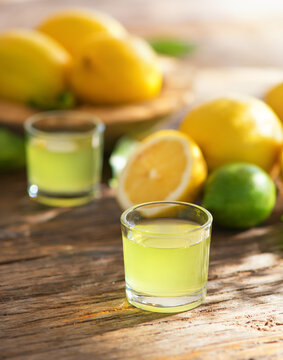Limoncello, Italian liqueur with lemons. Traditional Mediterranean sweet shot alcoholic drink close up, little glasses and fresh citrus fruits on wooden table. Vertical image