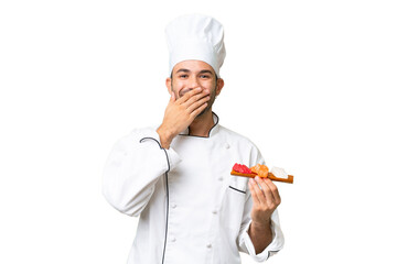 Young caucasian chef holding a sushi over isolated background happy and smiling covering mouth with hand