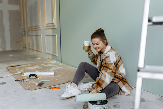 Smiling woman with paint roller having coffee in messy apartment