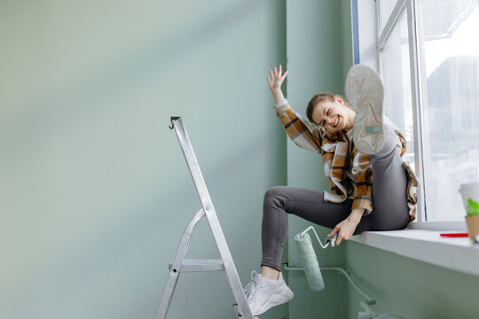 Cheerful young woman with paint roller on window sill at home