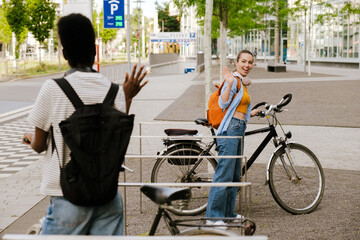 Multiracial women with bicycles waving hands to each other at city street