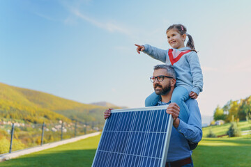 Father with his little daughter on piggyback, catching sun at solar panel,charging it at their...