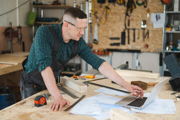 Portrait of a carpenter in protective glasses and work overalls uses a laptop in a workshop.