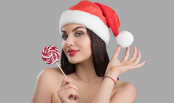 Christmas woman. Beauty winter model girl in Santa Claus hat with red lips and xmas lollipop candy in her hand. Joy. Surprised expression, smile. Closeup portrait over grey background