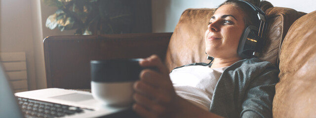 Pretty smiling girl listening to music from headphones with pleasure at coworking office, lying on sofa, using laptop, drinking hot coffe from nice cup and resting.