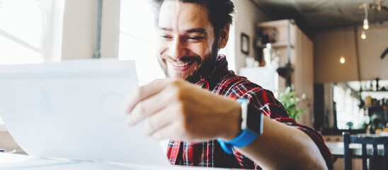 Young stubbled man working in big bright office space. Smiling and looking at the paper plan