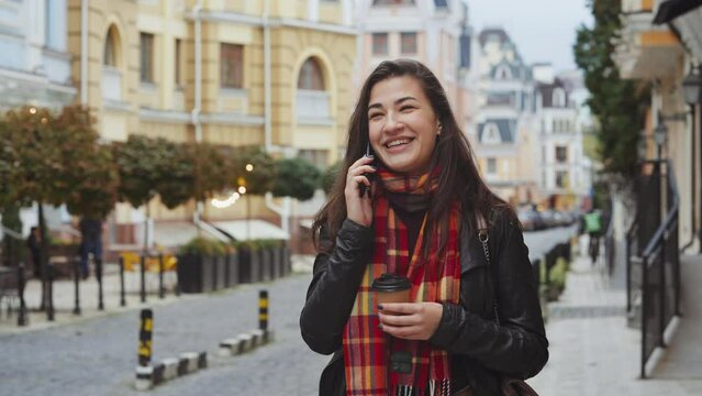Cheerful brunette woman with coffee cup wearing leather jacket with checkered scarf talking on phone, blurred old building background. Young female waiting for taxi in city. Concept of communication