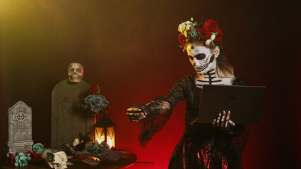Traditional goddess shopping online with credit card, using laptop to make electronic payment. Looking like lady of the holy death or santa muerte, paying internet transaction on pc.