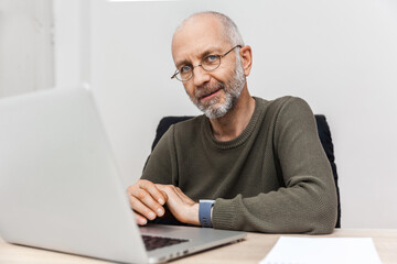 The old man works on his laptop and looks at the camera with a smile