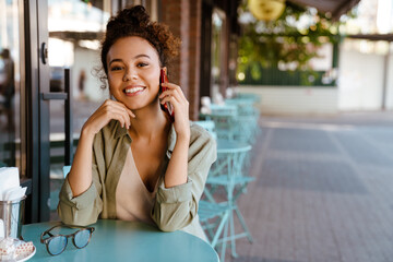 White young woman talking to cellphone while sitting in cafe outdoors